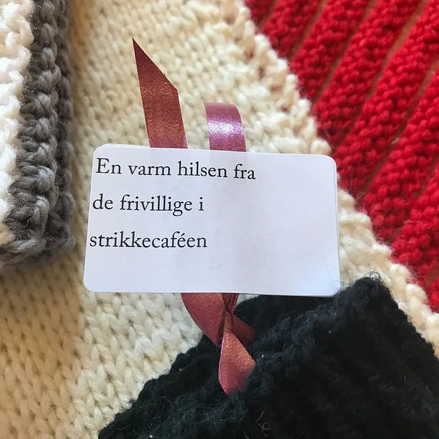 Et mærke med et bånd på strikkede ting, hvor der står "En varm hilsen fra de frivillige i strikkecaféen."