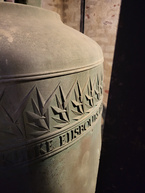 Nærbillede af en kirkeklokke med inskription og udskåret mønster.