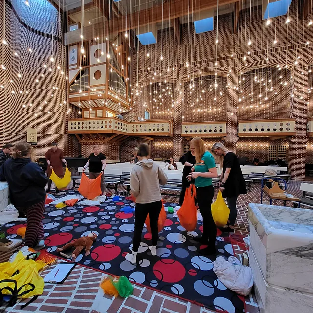 Folk rydder op i farverige poser og legetøj i en murstenskirke.